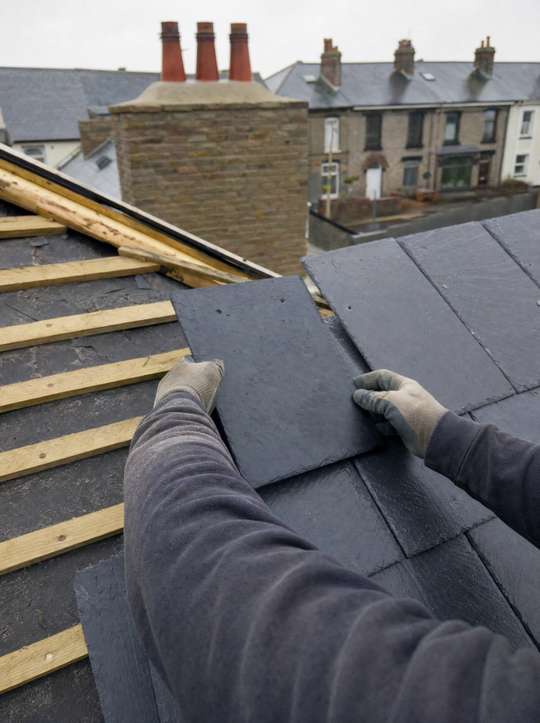 A roofer fitting a slate tile on a pitched roof above Birmingham rooftops.